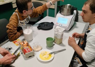 Dwaj mężczyźni przygotowują posiłek przy użyciu wielofunkcyjnego urządzenia kuchennego. Na stole są widoczne składniki potrzebne do stworzenia dania.