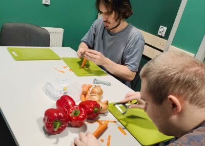 Dwaj mężczyźni siedząc przy stole, obierają marchewkę. Na stole ułożone są także inne warzywa.