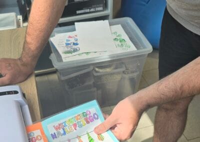 Uczestnik przy użyciu laminarki dokonuje zabezpieczenia laurek przygotowanych z okazji Dnia Ojca. Wykonując tą czynność pochyla się nad biurkiem, jego wzrok jest skierowany na urządzenie.