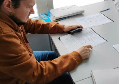 Uczestnik siedząc przy stole, wykonuje czynność dziurkowania kartki przy użyciu specjalnego artykułu biurowego.