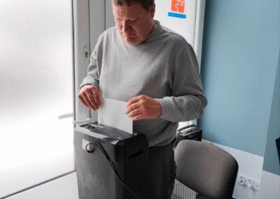 Mężczyzna niszczy kartkę papieru przy pomocy niszczarki. Spogląda w kierunku wspomnianego urządzenia.