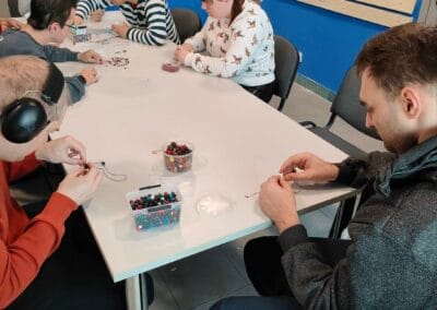 Pięć osób na zdjęciu nawleka ozdobne koraliki na sznureczki. Jeden mężczyzna przygląda się.