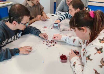 Pięć osób na zdjęciu podczas zajęć rękodzieła nawlekają ozdobne koraliki na sznureczki w celu zrobienia bransoletek.