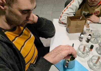 Dwóch mężczyzn siedzi przy stole. Jeden z nich wlewa olejek zapachowy do kubeczka z woskiem.