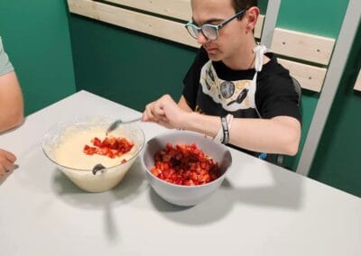 Uczestnik siedząc przy stole, przekłada owoce z jednej miski do drugiej z masą do babeczek.