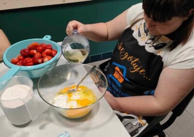 Uczestniczka wlewa olej do miski z innymi składnikami niezbędnymi do stworzenia babeczek. Na stole widoczne są truskawki, mąka oraz soda oczyszczona.
