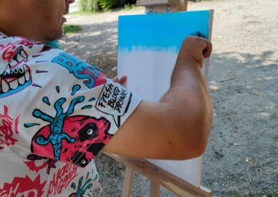 Mężczyzna stojąc w plenerze maluje wałkiem tło na płotnie. W oddali widać budynek.