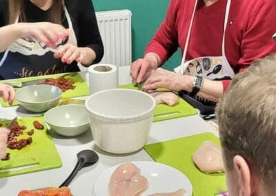 Trzy osoby przygotowują posiłek. Dwie z nich kroją mięso. Kobieta kroi suszone pomidory.