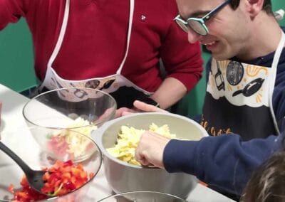 Uczestnik ŚDS podczas zajęć kulinarnych nakłada makaron w celu przygotowania sałatki. Zaproszony gość przygląda się.