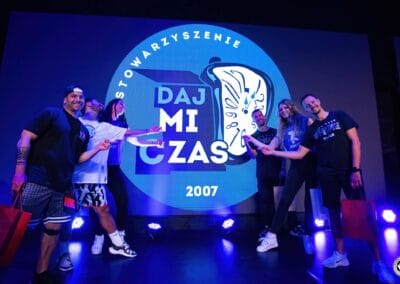 Sześć osób na scenie wskazuje rękami na logo stowarzyszenia "Daj mi czas".