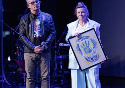 Mężczyzna i kobieta stoją na scenie podczas wydarzenia " Tanecznym krokiem w niebieski świat autyzmu". Kobieta trzyma w rękach obraz.