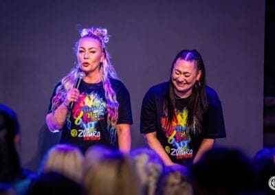 Dwie kobiety stoją na scenie podczas wydarzenia "Tanecznym krokiem w niebieski świat autyzmu". Jedna z nich zwraca się do publiczności przez mikrofon.