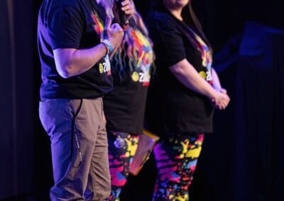 Trzy osoby stoją na scenie - mężczyzna (wypowiada się do publiczności używając mikrofonu) oraz dwie kobiety (jedna spoglądająca w kierunku publiczności, druga w stronę wypowiadającego się mężczyzny).