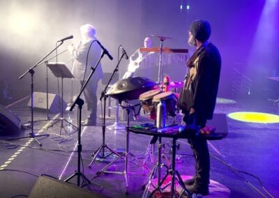 Trzech muzyków występuje na scenie, na której ustawione są instrumenty i sprzęt muzyczny.