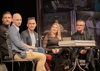 Czterech mężczyzn i jedna kobieta siedzą na krzesłach na scenie teatralnej.