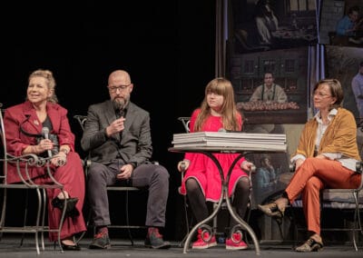 Cztery osoby siedzą na scenie na krzesłach: dwie kobiety, mężczyzna i dziewczynka. Jedna z kobiet trzyma w dłoni mikrofon.