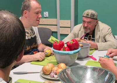 Trzech mężczyzn siedzi przy stole podczas zajęć kulinarnych. Dwóch z nich rozmawia, a trzeci obiera ziemniaki.