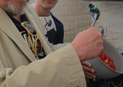 Dwóch mężczyzn pozuje do zdjęcia podczas zajęć kulinarnych. Jeden z nich wrzuca do garnka pomidory.