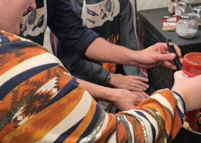 Kobieta wlewa passatę pomidorową do garnka. Dwóch mężczyzn przygląda się temu. Jeden z nich trzyma łopatkę kuchenną.