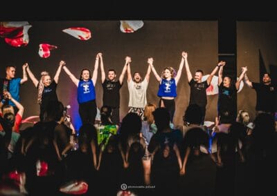 Instruktorzy zumby stoją na scenie trzymając się za ręce w górze. Skierowani są w stronę publiczności.