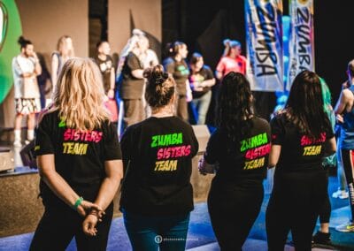 Cztery kobiety w koszulkach "Zumba sisters" stoją tyłem do obiektywu. Są zwrócone przodem do sceny.