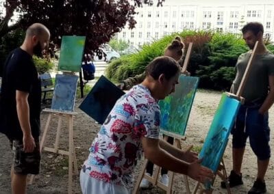 Cztery osoby są widoczne na zdjęciu. Trzy z nich ustawiają powstałe obrazy na sztalugach. Jeden mężczyzna stoi, przyglądając się tym działaniom.