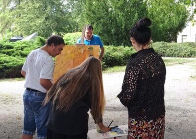 Trzy osoby pracują nad tworzeniem obrazu w plenerze. Czwarta stoi, będąc oddaloną od