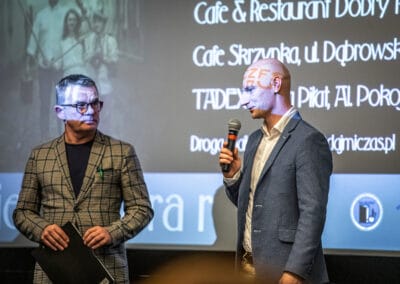 Na scenie stoi dwóch mężczyzn. Jeden z nich trzymając mikrofon, zwraca się w kierunku publiczności.