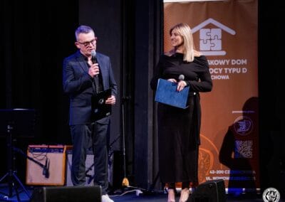 Fotografia przedstawia mężczyznę (Prezesa Stowarzyszenia Daj mi czas), który przy użyciu mikrofonu przemawia do publiczności. Drugą widoczną osobą jest Magdalena Woch - Dyrektor Teatru im. Adama Mickiewicza w Częstochowie. Obie osoby znajdują się na scenie teatralnej.