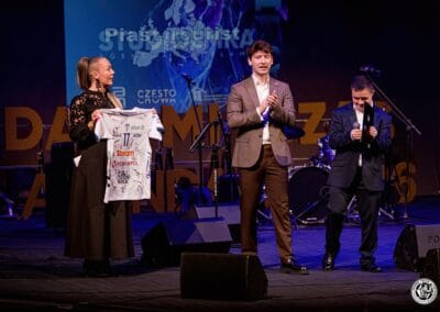 Fotografia przedstawia trzy osoby - dwóch mężczyzn prowadzących galę prezentacji kalendarza "Barwy Sportu okiem Skowronka" oraz kobietę, która asystuje przy licytacji oraz trzyma w dłoniach siatkarską koszulkę. Wszystkie z tych osób znajdują się na scenie teatralnej.