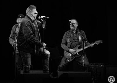 Na zdjęciu widoczni są muzycy - Muniek Staszczyk (wokalista), Krzysztof Niedźwiecki (gitarzysta) i saksofonista. Mężcyzźni znajdują się na scenie teatralnej.