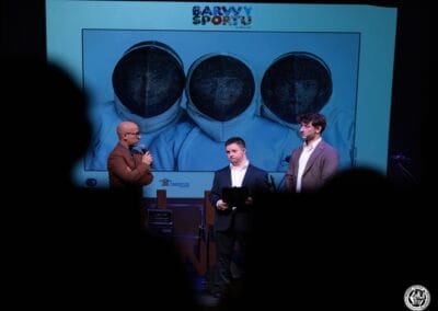 Na zdjęciu są widoczne trzy postacie - dwaj prowadzący galę prezentacji kalendarza "Barwy Sportu okiem Skowronka" oraz fotograf - Grzegorz Skowronek. W tle wyświetlane jest zdjęcie z wcześniej wspomnianego kalendarza.