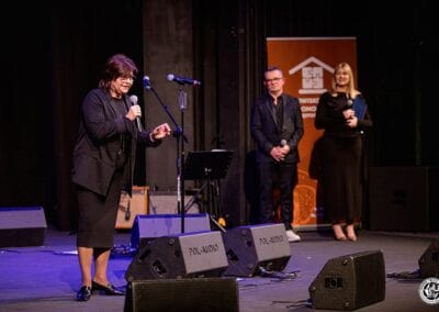 Na zdjęciu przedstawione są trzy postacie - na pierwszym planie widoczna jest kobieta (Izabela Leszczyna - posłanka na Sejm RP), która zwraca się do publiczności, wypowiadając się do mikrofonu. W tle widoczny jest mężczyzna - Prezes Stowarzyszenia Daj mi czas - oraz kobieta - Magdalena Woch (dyrektor Teatru im. Adama Mickiewicza w Częstochowie). Wszystkie postacie znajdują się na scenie teatralnej.