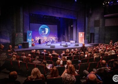 Na zdjęciu przedstawiona jest sala teatralna. Na widowni zasiadają goście. Scena jest oświetlona, ale w przedstawionym momencie - pusta. Na scenie widoczne są m.in. mikrofony, instrumenty muzyczne, banery Stowarzyszenia Daj mi czas oraz ŚDS typu D.