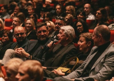 Na zdjęciu widoczny jest mężczyzna wypowiadający się do mikrofonu, siedzący wśród publiczności. Część osób kieruje swój wzrok na wypowiadającego się.