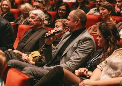 Na pierwszym planie zdjęcia widoczny jest mężczyzna siedzący wśród publiczności, wypowiadający się do mikrofonu.