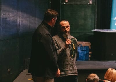 Na fotografii widoczni są dwaj mężczyźni - jeden wypowiada się przy użyciu mikrofonu, drugi kieruje swoją uwagę na przemawiającego.