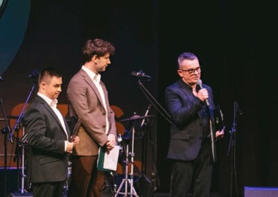 Na fotografii widocznych jest trzech mężczyzn - dwaj prowadzący galę prezentacji kalendarza "Barwy Sportu okiem Skowronka" oraz prezes Stowarzyszenia Daj mi czas, który zwraca się do publiczności, trzymając w ręku mikrofon. Wszystkie osoby znajdują się na scenie teatralnej.