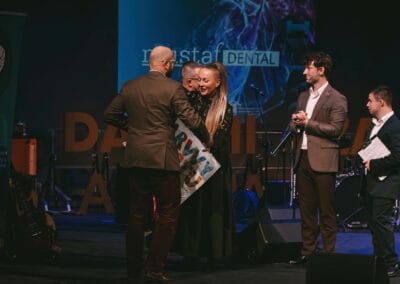 Na zdjęciu widocznych jest pięć postaci - dwaj prowadzący galę prezentacji kalendarza "Barwy Sportu okiem Skowronka", prezes Stowarzyszenia Daj mi czas oraz kobieta, która wręcza mężczyźnie wylicytowany przedmiot. Wszystkie osoby znajdują się na scenie.