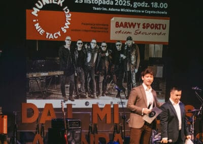 Na fotografii przedstawieni są dwaj mężczyźni - prowadzący galę prezentacji kalendarza "Barwy Sportu okiem Skowronka". Na drugim planie widoczny jest plakat (wyświetlony na ekranie), który zapowiada koncert Muńka i NieTakichŚwiętych.