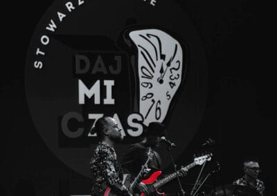 Na fotografii przedstawieni są muzycy grający na instrumentach takich jak gitara czy perkusja. W tle widoczne jest logo Stowarzyszenia Daj mi czas. Mężczyźni występują na scenie.