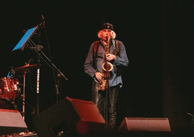 Na fotografii przedstawiony jest muzyk grający na saksofonie. Postać znajduje się na scenie.