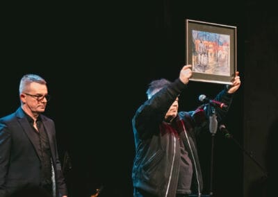 Na zdjęciu przedstawieni są dwaj mężczyźni - Muniek Staszczyk oraz prezes Stowarzyszenia Daj mi czas. Muzyk trzyma w dłoniach obramowany rysunek i przedstawia go w kierunku publiczności.